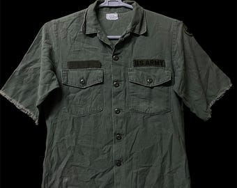 Könnte beinhalten: Ein olivgrünes Kurzarmhemd mit Kragen, zwei Brusttaschen mit Knopfklappen und einem schwarzen Aufnäher mit der in Weiß gestickten Aufschrift U.S. ARMY. Die Ärmel haben ausgefranste Kanten und einen Aufnäher an der Schulter.