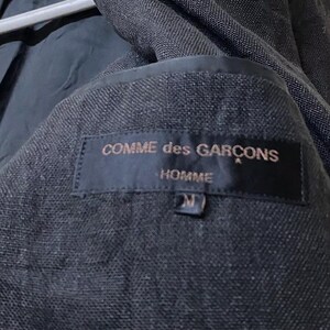 May include: Close-up of the interior of a dark gray blazer. A black label is sewn inside, with the words "COMME des GARCONS HOMME" in gold lettering. The size "M" is also visible on the label.