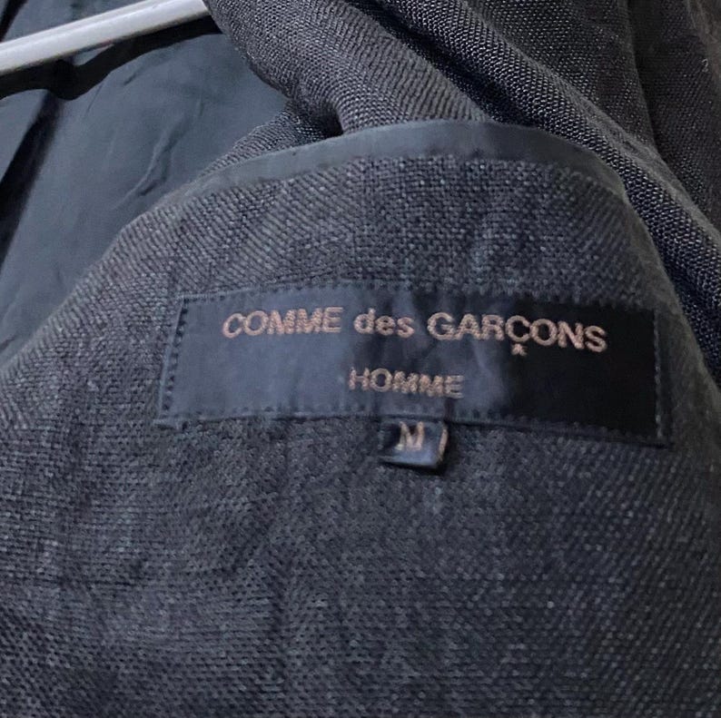 May include: Close-up of the interior of a dark gray blazer. A black label is sewn inside, with the words "COMME des GARCONS HOMME" in gold lettering. The size "M" is also visible on the label.