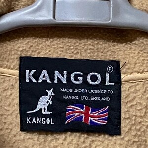 May include: Close-up of a tan fabric with a black label. The label features the white text "KANGOL" above a kangaroo logo and the text "MADE UNDER LICENCE TO KANGOL LTD., ENGLAND" with a Union Jack flag.