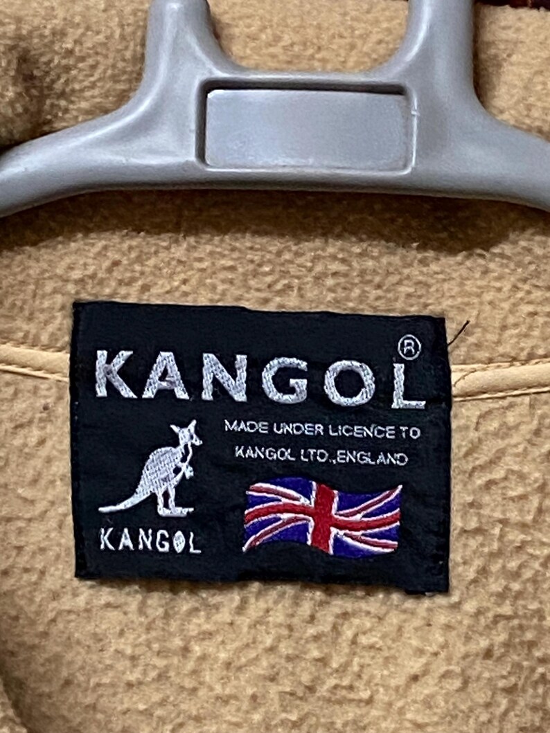 May include: Close-up of a tan fabric with a black label. The label features the white text "KANGOL" above a kangaroo logo and the text "MADE UNDER LICENCE TO KANGOL LTD., ENGLAND" with a Union Jack flag.