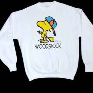 Peut inclure: Sweat-shirt blanc avec un personnage d'oiseau de dessin animé jaune, Woodstock, portant une casquette bleue et orange. Le mot "WOODSTOCK" est imprimé en noir sous le personnage. Le sweat-shirt a des manches longues et un col rond.