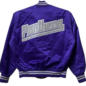 Puede incluir: Una chaqueta bomber de satén morado con un logotipo "Panthers" blanco y gris en la espalda. La chaqueta tiene un cuello, puños y cintura de punto de canalé blanco y gris.