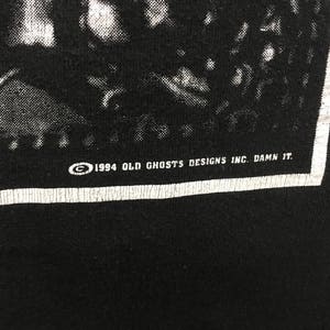 May include: Black t-shirt with a white border and text. The text reads "&copy; 1994 OLD GHOSTS DESIGNS INC. DAMN IT." The image is a close-up of the bottom right corner of the shirt.