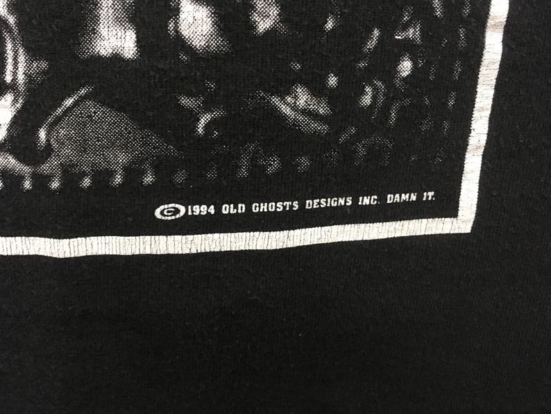 May include: Black t-shirt with a white border and text. The text reads "&copy; 1994 OLD GHOSTS DESIGNS INC. DAMN IT." The image is a close-up of the bottom right corner of the shirt.