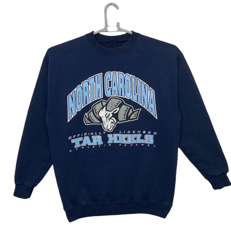 May include: A navy blue sweatshirt with a light blue and gray graphic of a ram's head with the text "North Carolina" and "Tar Heels" in a white arch above and below the ram's head. The text "Officially Licensed Athletic Apparel" is printed below the ram's head.