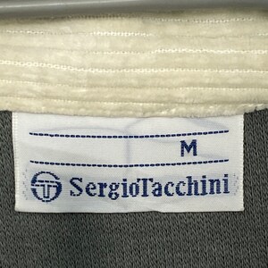 May include: A white tag with black stitching and the text "Sergio Tacchini" in blue lettering. The tag also has a small logo of a circle with a "T" inside. The tag is attached to a gray piece of clothing and has the letter "M" printed on it.