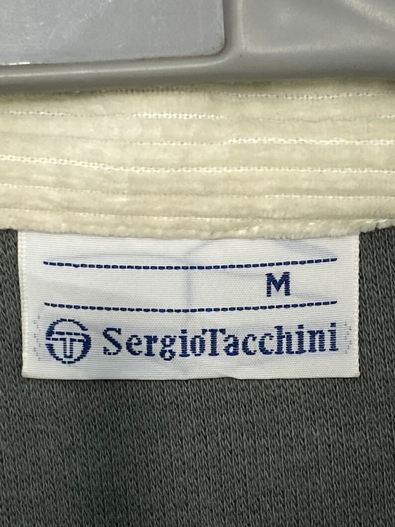 May include: A white tag with black stitching and the text "Sergio Tacchini" in blue lettering. The tag also has a small logo of a circle with a "T" inside. The tag is attached to a gray piece of clothing and has the letter "M" printed on it.