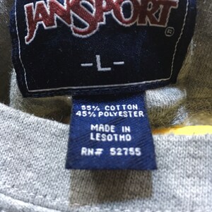 Puede incluir: Una etiqueta azul y blanca con el texto "JANSPORT" en letras rojas, "L" en letras blancas, "55% ALGOD&Oacute;N 45% POLI&Eacute;STER", "HECHO EN LESOTHO" y "RN# 52755".