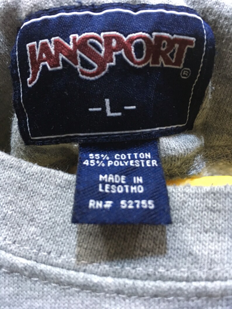 Puede incluir: Una etiqueta azul y blanca con el texto "JANSPORT" en letras rojas, "L" en letras blancas, "55% ALGOD&Oacute;N 45% POLI&Eacute;STER", "HECHO EN LESOTHO" y "RN# 52755".