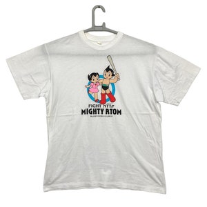 Op de afbeelding: Wit T-shirt met een afbeelding van Astro Boy en zijn zus die een honkbalknuppel vasthouden. De tekst "Fight NTT!! Mighty Atom" staat onder de afbeelding.