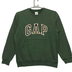 Puede incluir: Una sudadera verde de cuello redondo con un gran logotipo "GAP" bordado en letras azul marino y doradas.