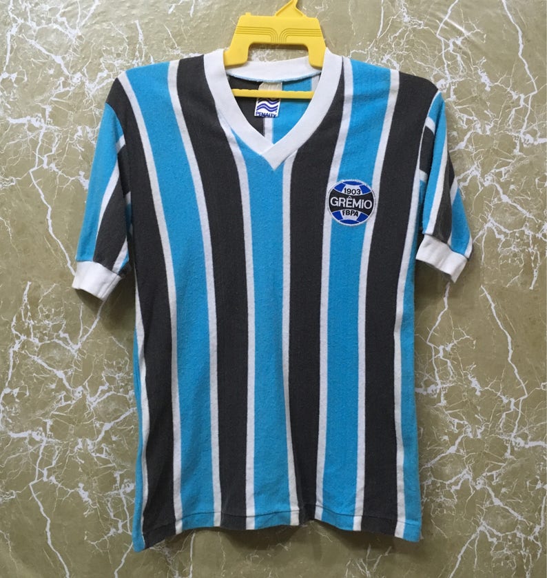 May include: A blue and black striped football shirt with a white v-neck collar. The shirt has a patch on the chest that says "1903 GREMIO FBPA".