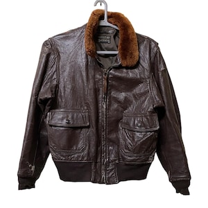 May include: A brown leather bomber jacket with a shearling collar. The jacket has two pockets on the front and a zip closure.