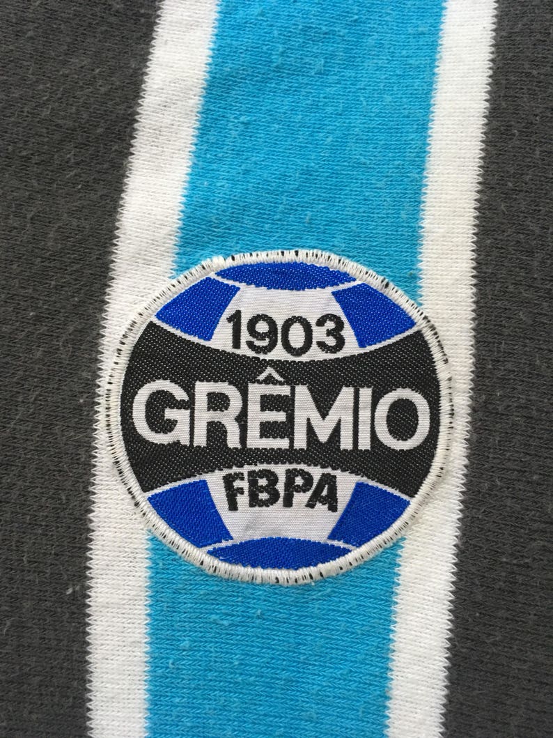 May include: A black and white circular patch with blue stitching. The patch features a football with the text "1903" above it, "GREMIO" in a large font, and "FBPA" below the football.