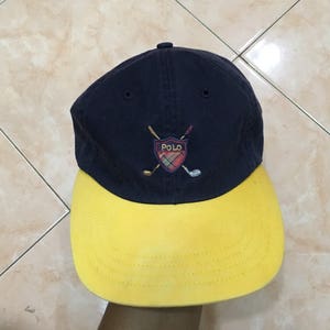 May include: A navy blue and yellow baseball cap with a logo of a shield with golf clubs and the word "POLO" inside.