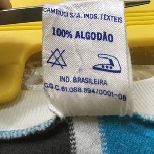 May include: White clothing label with blue text that reads "CAMBUCI S/A. INDS. TEXTEIS 100% ALGOD&Atilde;O IND. BRASILEIRA C.G.C. 61.088.894/0001-08". The label has a blue triangle with an X inside and a blue iron with three dots inside.