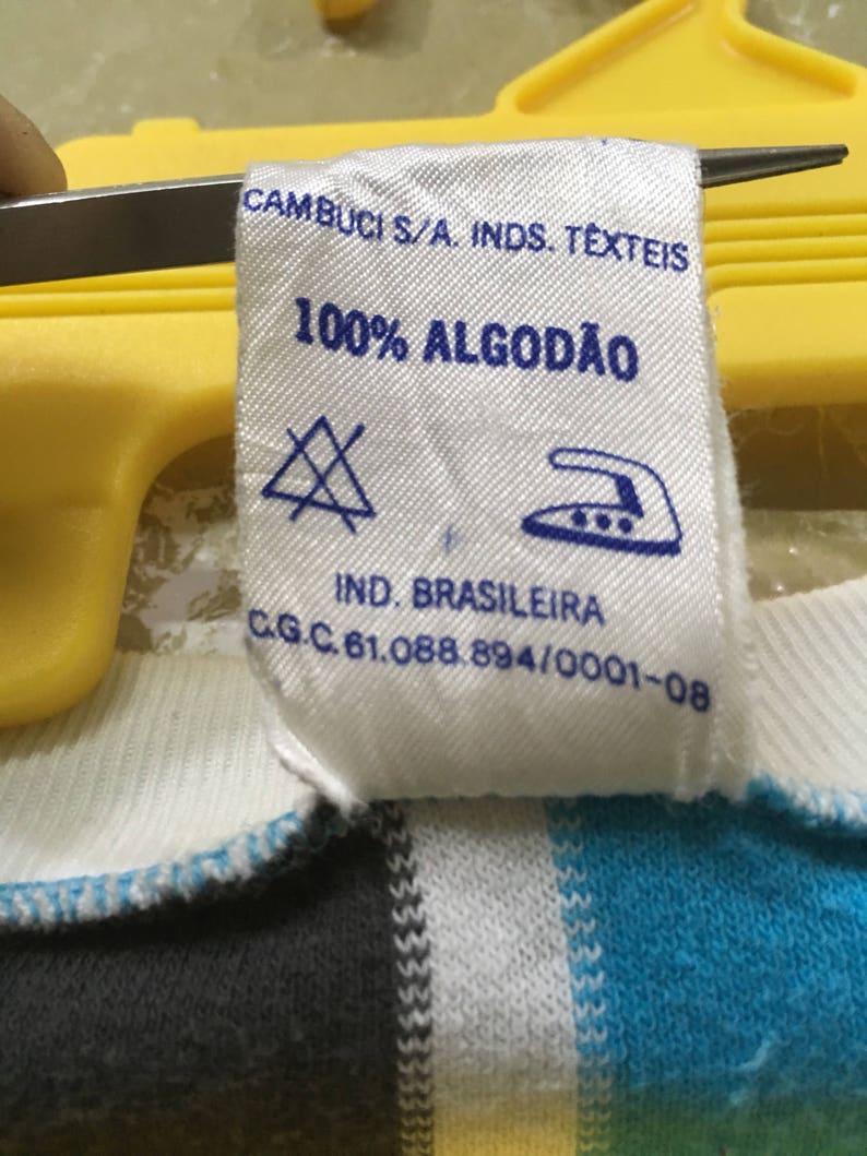 May include: White clothing label with blue text that reads "CAMBUCI S/A. INDS. TEXTEIS 100% ALGOD&Atilde;O IND. BRASILEIRA C.G.C. 61.088.894/0001-08". The label has a blue triangle with an X inside and a blue iron with three dots inside.