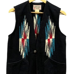 Könnte beinhalten: Schwarze Wollweste mit einem Design im Südwest-Stil. Die Weste hat ein geometrisches Muster in Rot, Weiß und Blau. Sie hat zwei Vordertaschen und einen einzelnen Knopfverschluss. Sie hängt an einem Holzkleiderbügel.