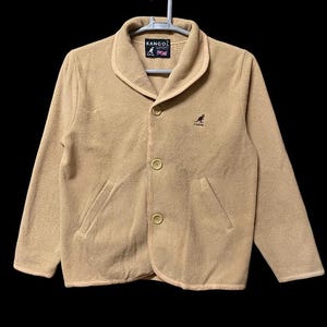 May include: Tan fleece jacket with a shawl collar, two front pockets, and three buttons. The jacket features the "KANGOL" logo on the label and a kangaroo logo on the chest. The jacket is hanging on a white hanger.