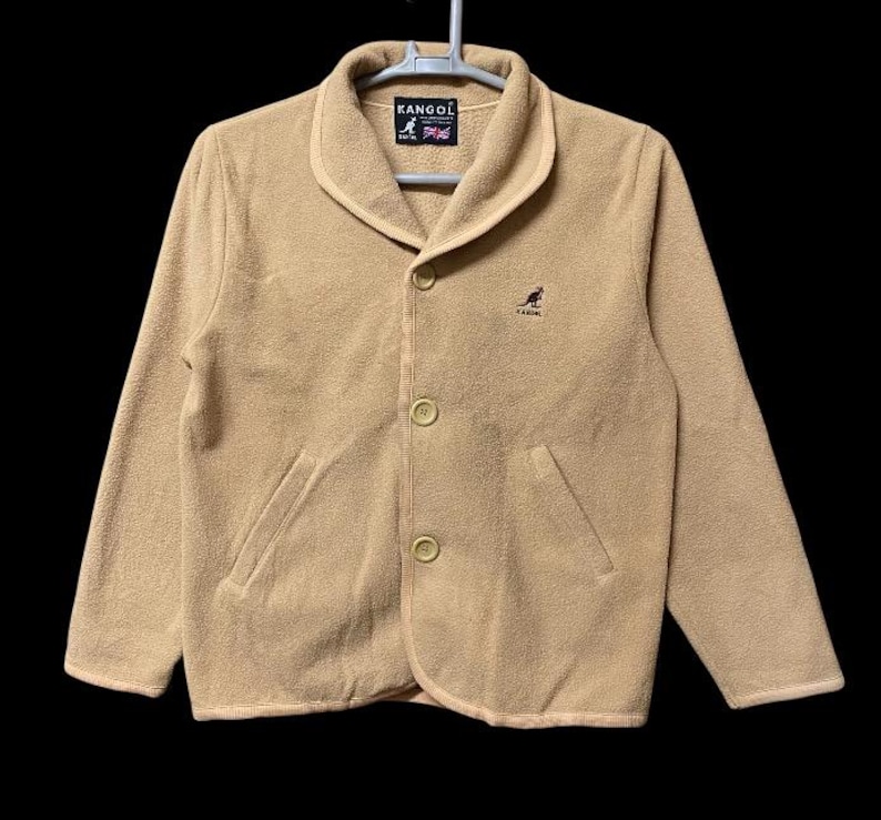 May include: Tan fleece jacket with a shawl collar, two front pockets, and three buttons. The jacket features the "KANGOL" logo on the label and a kangaroo logo on the chest. The jacket is hanging on a white hanger.