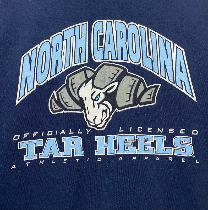 May include: A navy blue t-shirt with a light blue graphic of the North Carolina Tar Heels logo. The logo features a ram's head with a blue eye and the text "Officially Licensed Tar Heels Athletic Apparel".
