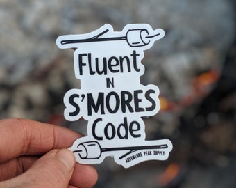 Könnte beinhalten: Weißer Aufkleber mit schwarzem Text und Illustrationen eines Stocks mit einer Marshmallow an jedem Ende. Der Text lautet "Fluent in S'mores Code" und "Adventure Peak Supply".