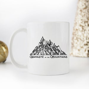 K&ouml;nnte beinhalten: Wei&szlig;e Keramik-Tasse mit einer schwarz-wei&szlig;en Bergketten-Illustration und dem Text "Namast&eacute; in the Mountains". Die Tasse hat einen gebogenen Henkel und eine gl&auml;nzende Oberfl&auml;che.