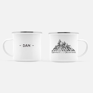K&ouml;nnte beinhalten: Zwei wei&szlig;e Emaille-Tassen mit silbernen R&auml;ndern. Eine Tasse hat den Namen "- DAN -" in Braun aufgedruckt. Die andere Tasse zeigt eine Schwarz-Wei&szlig;-Bergillustration mit dem Text "Namaste in the Mountains".