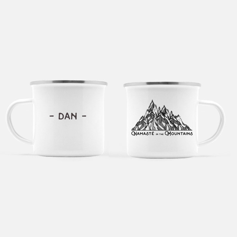 K&ouml;nnte beinhalten: Zwei wei&szlig;e Emaille-Tassen mit silbernen R&auml;ndern. Eine Tasse hat den Namen "- DAN -" in Braun aufgedruckt. Die andere Tasse zeigt eine Schwarz-Wei&szlig;-Bergillustration mit dem Text "Namaste in the Mountains".