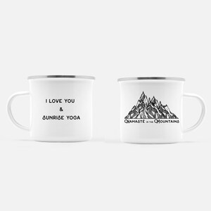K&ouml;nnte beinhalten: Zwei wei&szlig;e Emaille-Tassen mit silbernen R&auml;ndern. Eine Tasse hat den Text "I LOVE YOU & SUNRISE YOGA" in Schwarz. Die andere Tasse zeigt eine Schwarz-Wei&szlig;-Bergillustration mit dem Text "NAMAST&Eacute; in the MOUNTAINS".
