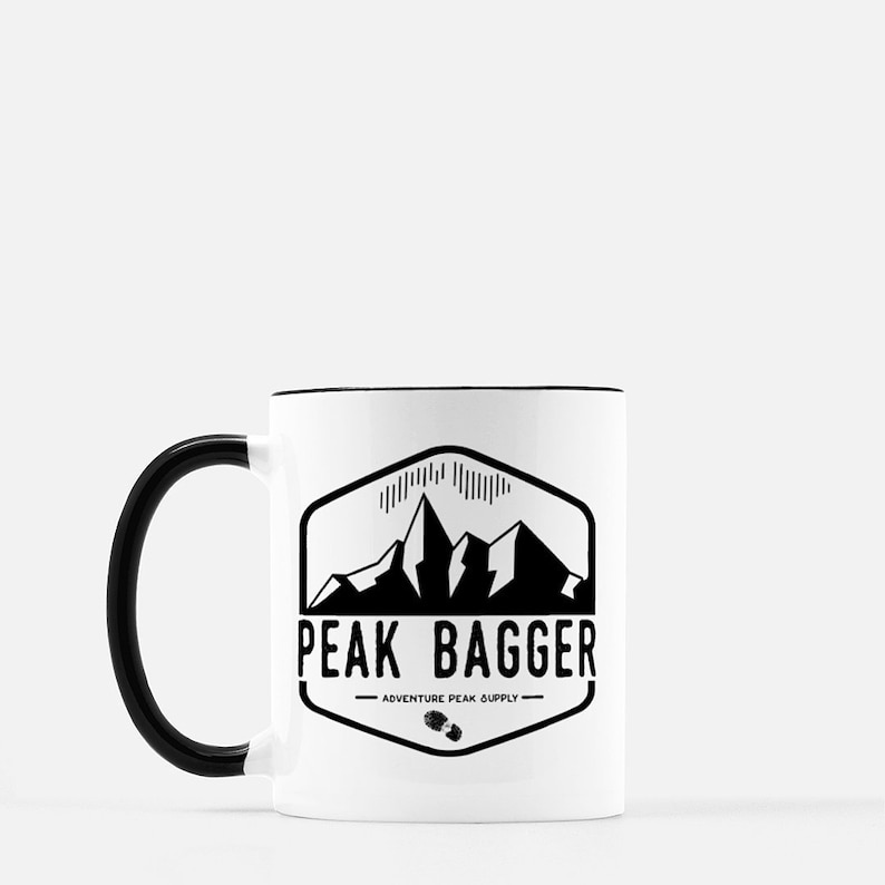 K&ouml;nnte beinhalten: Eine wei&szlig;e Keramiktasse mit schwarzem Henkel. Die Tasse zeigt eine schwarz-wei&szlig;e Grafik einer Bergkette mit dem Text "Peak Bagger - Adventure Peak Supply".