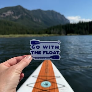 Puede incluir: Una pegatina blanca con texto azul y un gráfico de pala azul que dice "Go With The Float" y "Adventure Peak Supply". La pegatina se sostiene frente a una tabla de paddle surf en un lago con montañas en el fondo.