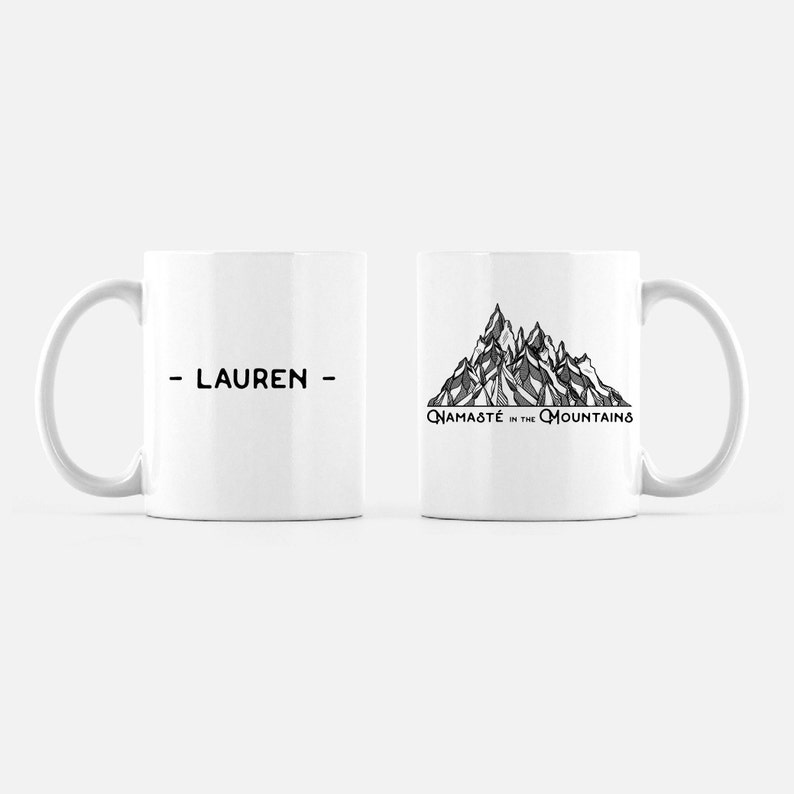 K&ouml;nnte beinhalten: Zwei wei&szlig;e Keramikbecher. Auf einem Becher ist der Name "- LAUREN -" in Schwarz aufgedruckt. Der andere Becher zeigt eine Schwarz-Wei&szlig;-Bergillustration mit dem Text "Namaste in the Mountains".