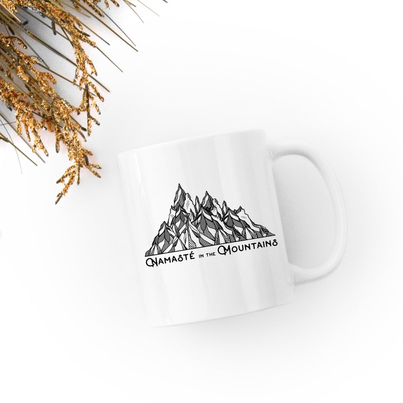 K&ouml;nnte beinhalten: Wei&szlig;e Keramiktasse mit einer schwarz-grauen Berglandschaftsillustration und dem Text "Namaste in the Mountains". Die Tasse hat einen gebogenen Henkel und ist f&uuml;r hei&szlig;e oder kalte Getr&auml;nke geeignet.
