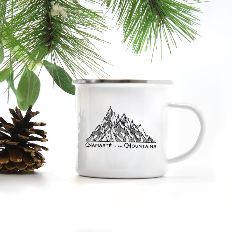 K&ouml;nnte beinhalten: Wei&szlig;er Emaillebecher mit silbernem Rand und Henkel. Der Becher zeigt eine schwarz-graue Bergkette mit dem Text "Namast&eacute; in the Mountains" in schwarzer Schrift. Eine Tannenzapfen und Gr&uuml;nzeug befinden sich im Hintergrund.