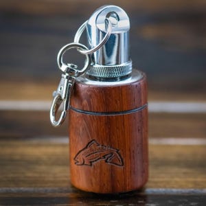 May include: A wooden keychain with a silver metal top and accents. The cylindrical body is made of dark brown wood and features a carved fish design. A silver keyring and clasp are attached.