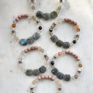 Puede incluir: Seis pulseras de piedras preciosas con cuentas de madera y piedras de labradorita cuadradas. Las pulseras presentan cuarzo transparente y otras cuentas coloridas, ensartadas en una banda elástica. Las pulseras se exhiben sobre una superficie de mármol blanco.