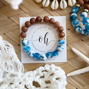 May include: A wooden bead bracelet with blue and white marbled beads. The bracelet is on a white card with the text "The Oil Haven Boutique" and "Oh" in a cursive font. The card also says "Aromatherapy Jewelry - Natural Products".