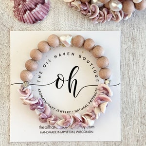 Pode incluir: Um bracelete de contas de madeira com conchas rosa e brancas, um fecho de coração branco e detalhes dourados. O bracelete está sobre um cartão branco com o texto "The Oil Haven Boutique, Joias de aromaterapia, Produtos naturais, theoilhavenboutique.com, Feito à mão em Appleton, Wisconsin".