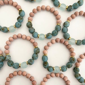 Pode incluir: Um conjunto de oito pulseiras com uma mistura de contas de madeira marrom claro e contas de vidro azul esverdeado com detalhes dourados. Cada pulseira tem uma única conta de vidro azul claro maior com detalhes dourados.