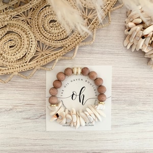 Pode incluir: Um bracelete de contas de madeira com pingentes de concha branca. O bracelete está em um cartão branco com o texto "The Oil Haven Boutique" e "oh" em uma fonte de escrita manuscrita.