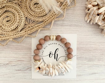 Può includere: Un braccialetto di perline di legno con ciondoli di conchiglia bianca. Il braccialetto è su una carta bianca con il testo "The Oil Haven Boutique" e "oh" in un font corsivo.