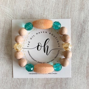 May include: Aromatherapy bracelet featuring light wood beads, peach-colored oval beads, and teal and yellow faceted beads. The bracelet is displayed on a white card with the text "THE OIL HAVEN BOUTIQUE" and "oh" logo.