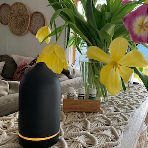 Puede incluir: Un difusor de aceites esenciales negro con una base brillante est&aacute; sobre una mesa de madera. Un jarr&oacute;n de tulipanes amarillos y morados est&aacute; detr&aacute;s del difusor. Tres botellas peque&ntilde;as de aceites esenciales est&aacute;n en un soporte de madera. Un camino de mesa de macram&eacute; est&aacute; sobre la mesa.