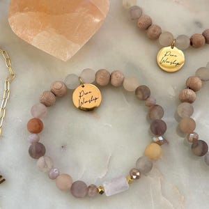 Może przedstawiać: Kilka bransoletek z koralików z różnymi koralikami w neutralnych odcieniach, w tym drewno, kwarc i szkło. Każda bransoletka ma złoty charms ze słowami "Pure Worship". Widoczny jest również kamień w kształcie serca.