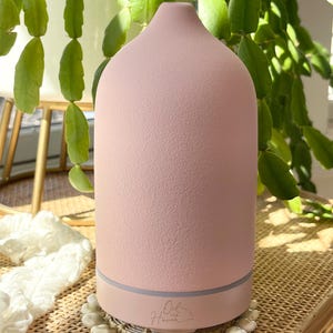 Puede incluir: Un difusor de aceites esenciales rosa con una superficie texturizada y una parte superior redondeada. La base es de color rosa claro con una banda gris. El difusor se encuentra sobre una esterilla tejida rodeada de peque&ntilde;os objetos blancos con forma de concha. El texto "Oil of Haven" es visible.