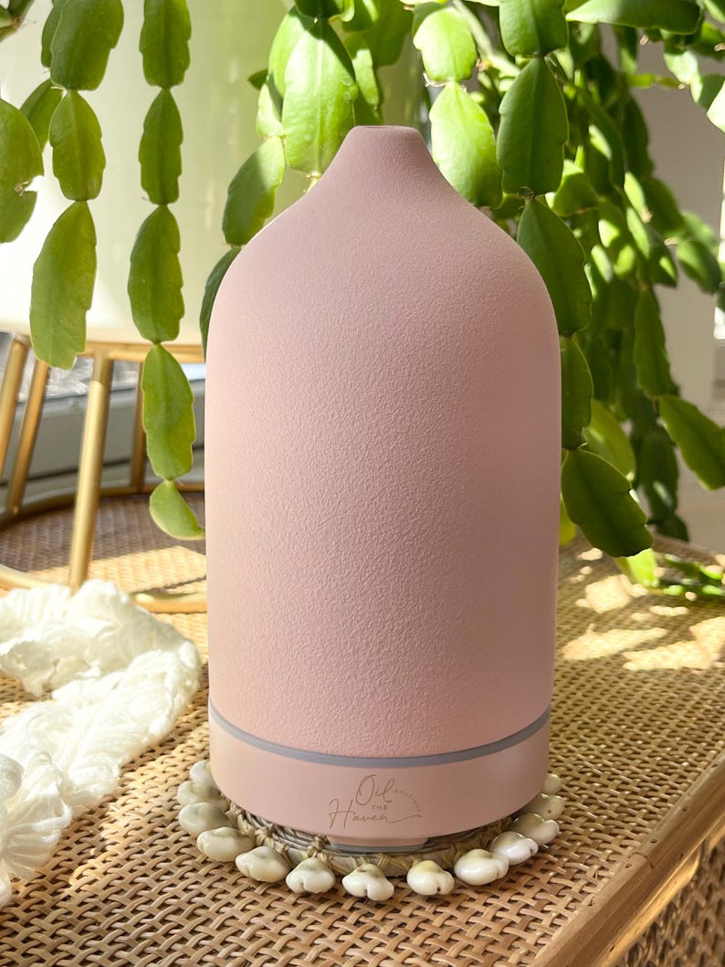 Puede incluir: Un difusor de aceites esenciales rosa con una superficie texturizada y una parte superior redondeada. La base es de color rosa claro con una banda gris. El difusor se encuentra sobre una esterilla tejida rodeada de peque&ntilde;os objetos blancos con forma de concha. El texto "Oil of Haven" es visible.