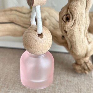May include: A small, round, frosted pink glass bottle suspended by a white cord. The bottle features a natural light-colored wooden cap with a hole for the cord, and a small wooden bead slider above the cap to adjust the length of the hanging loop.
