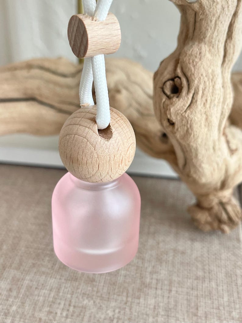 May include: A small, round, frosted pink glass bottle suspended by a white cord. The bottle features a natural light-colored wooden cap with a hole for the cord, and a small wooden bead slider above the cap to adjust the length of the hanging loop.